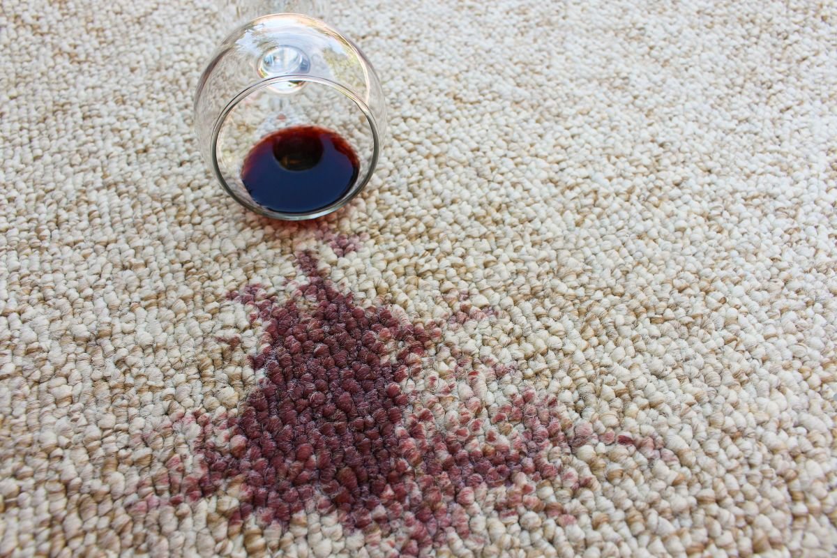 A glass of red wine tipped over on the floor, leaving a noticeable stain on the carpet, indicating a need for cleaning.